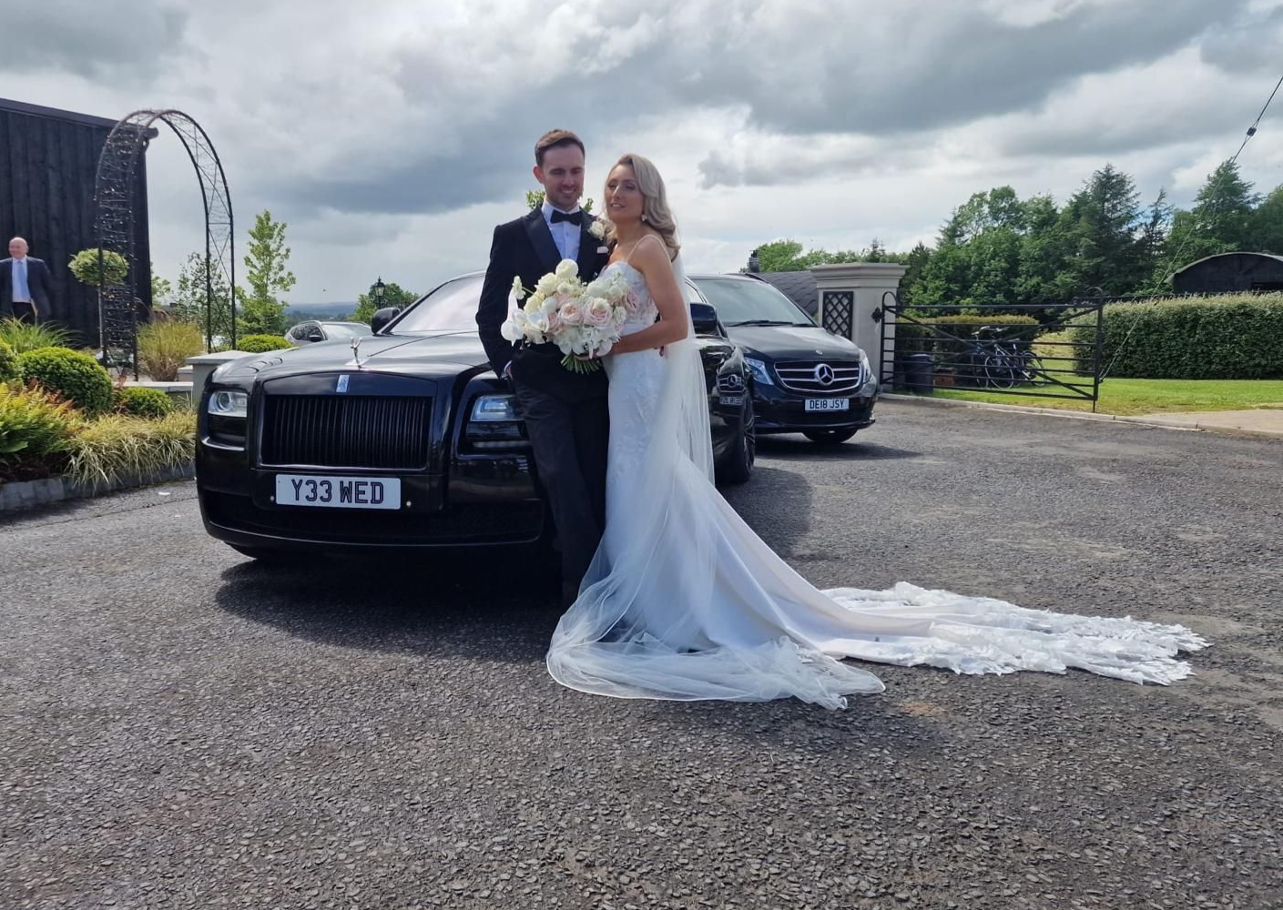 Bride and groom posing by a black car in a driveway, bride holding a bouquet and wearing a long white gown