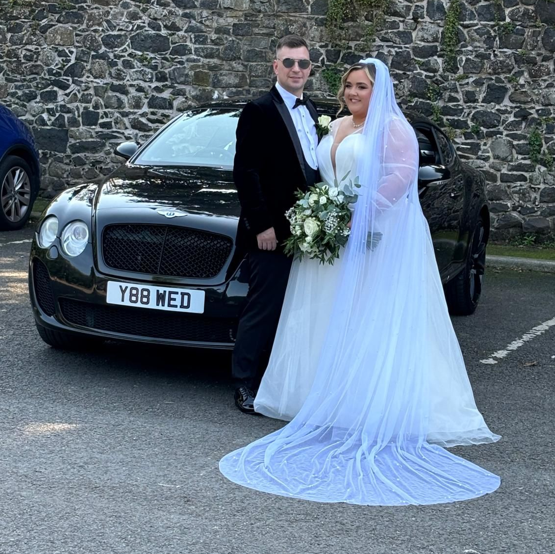 Bride and groom posing in front of a black luxury car against a stone wall