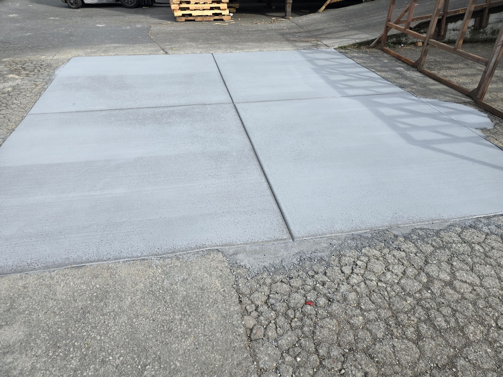 Concrete driveway and cracked asphalt transition near a curb and pallets in an outdoor lot