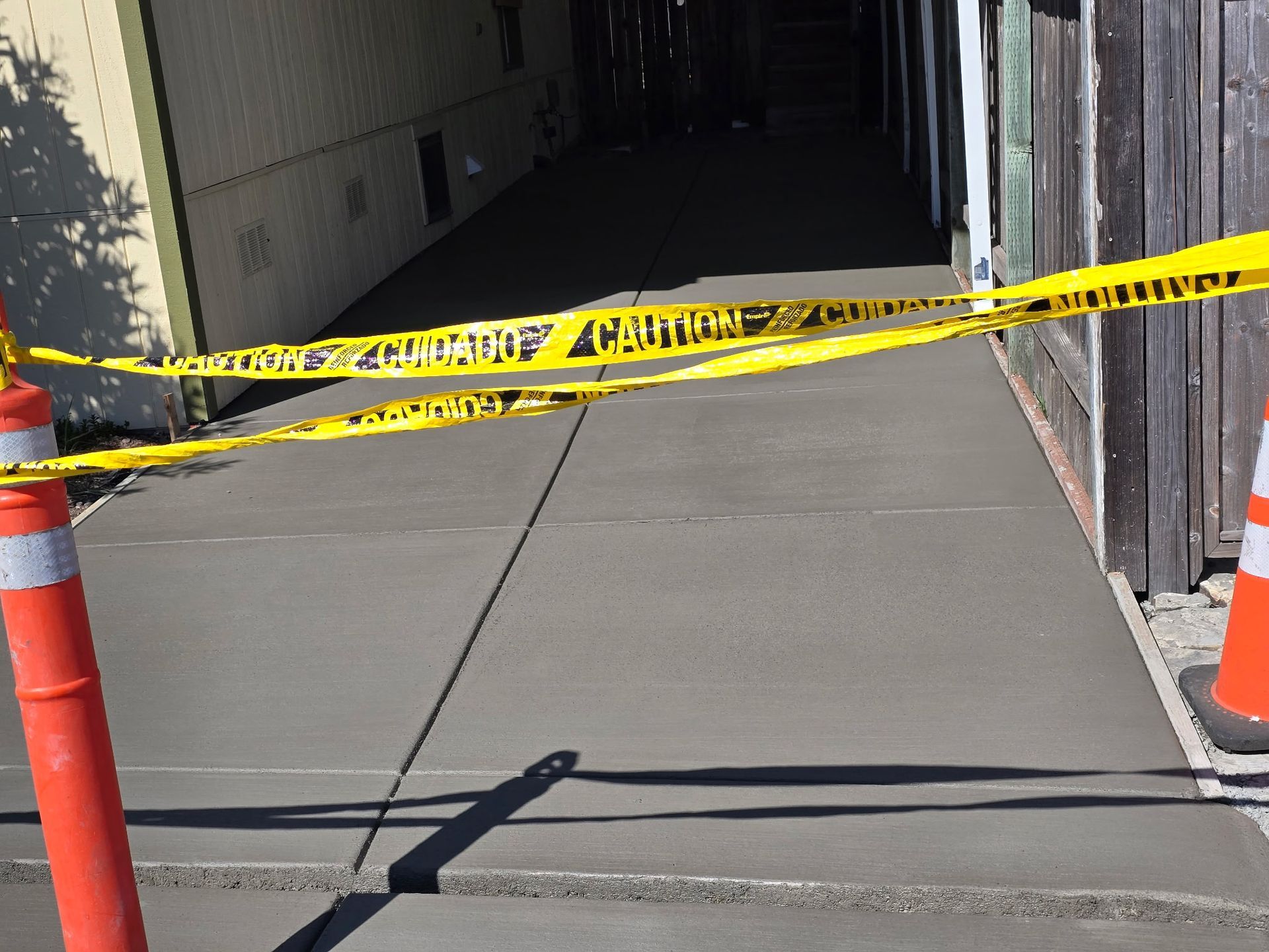 Yellow caution tape blocks a concrete walkway between buildings, with orange cones at the sides.