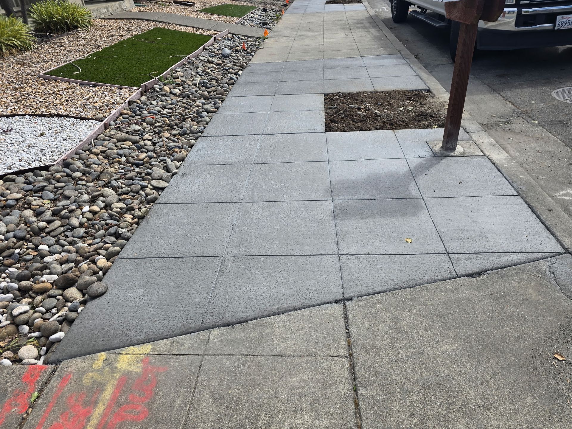 Sidewalk with gray paving slabs beside a rock garden and parked vehicle; one slab is lifted near a pole.