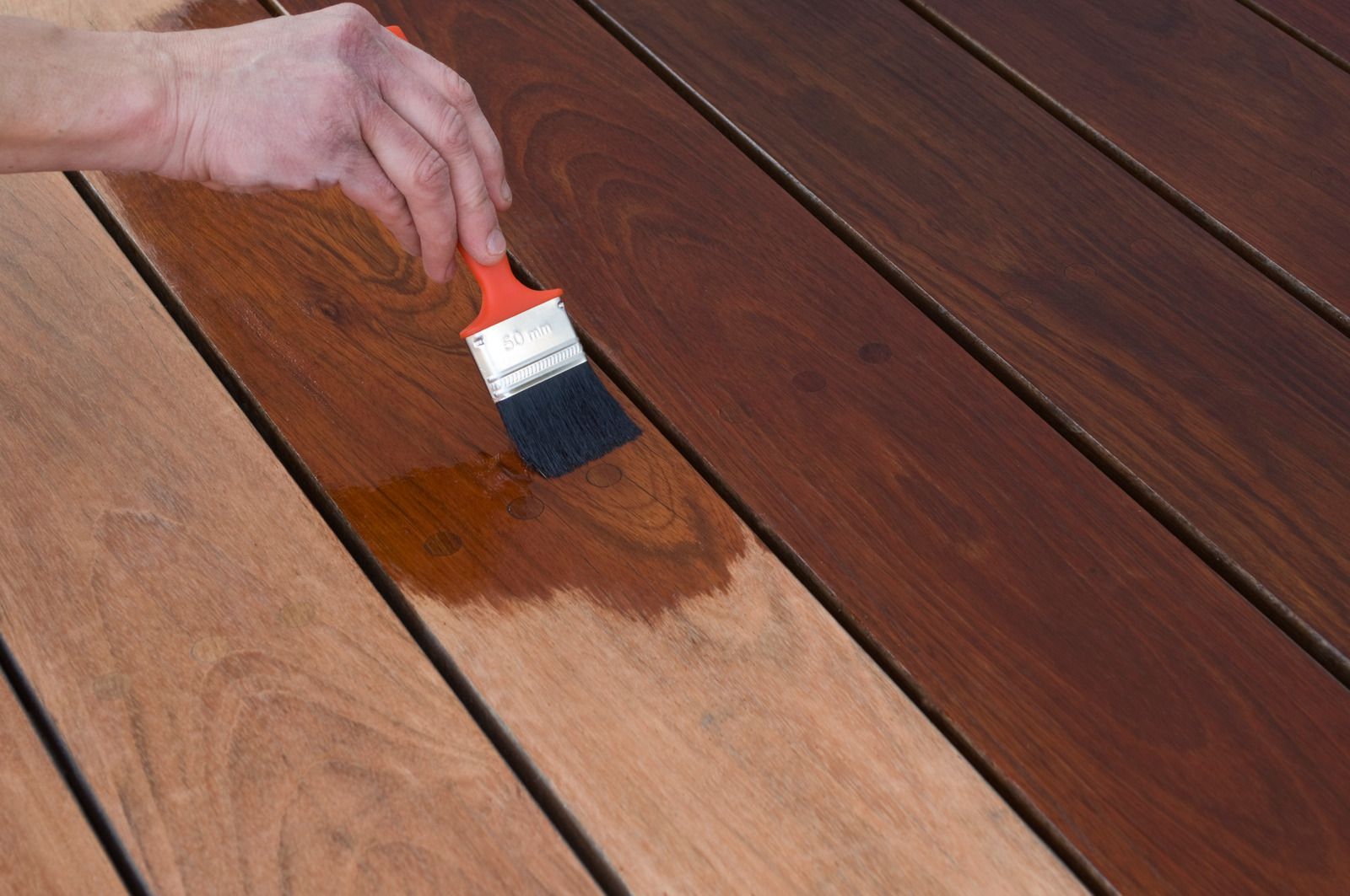 Hand applying brown stain to wooden deck planks with a brush.