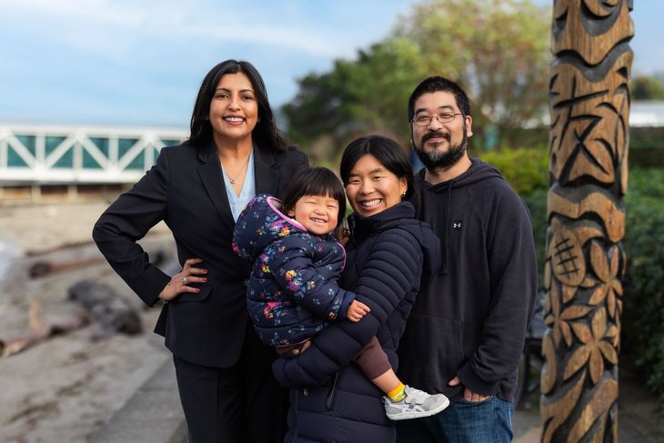 Four people smile outdoors near a waterfront, with a child held in front and carved totem pole beside them.