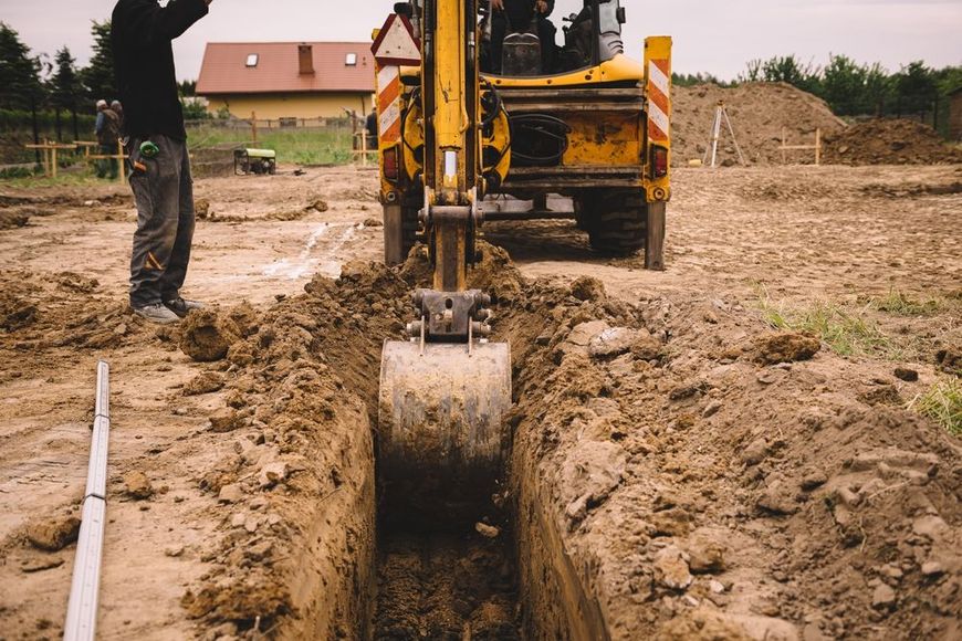 Yellow excavator digging a trench in brown dirt, a worker stands by.