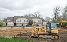 Bulldozer leveling dirt in front of a multi-unit house on a cloudy day.