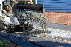 Skid steer pours gravel onto a black fabric-covered area, next to a blue house and brick wall.