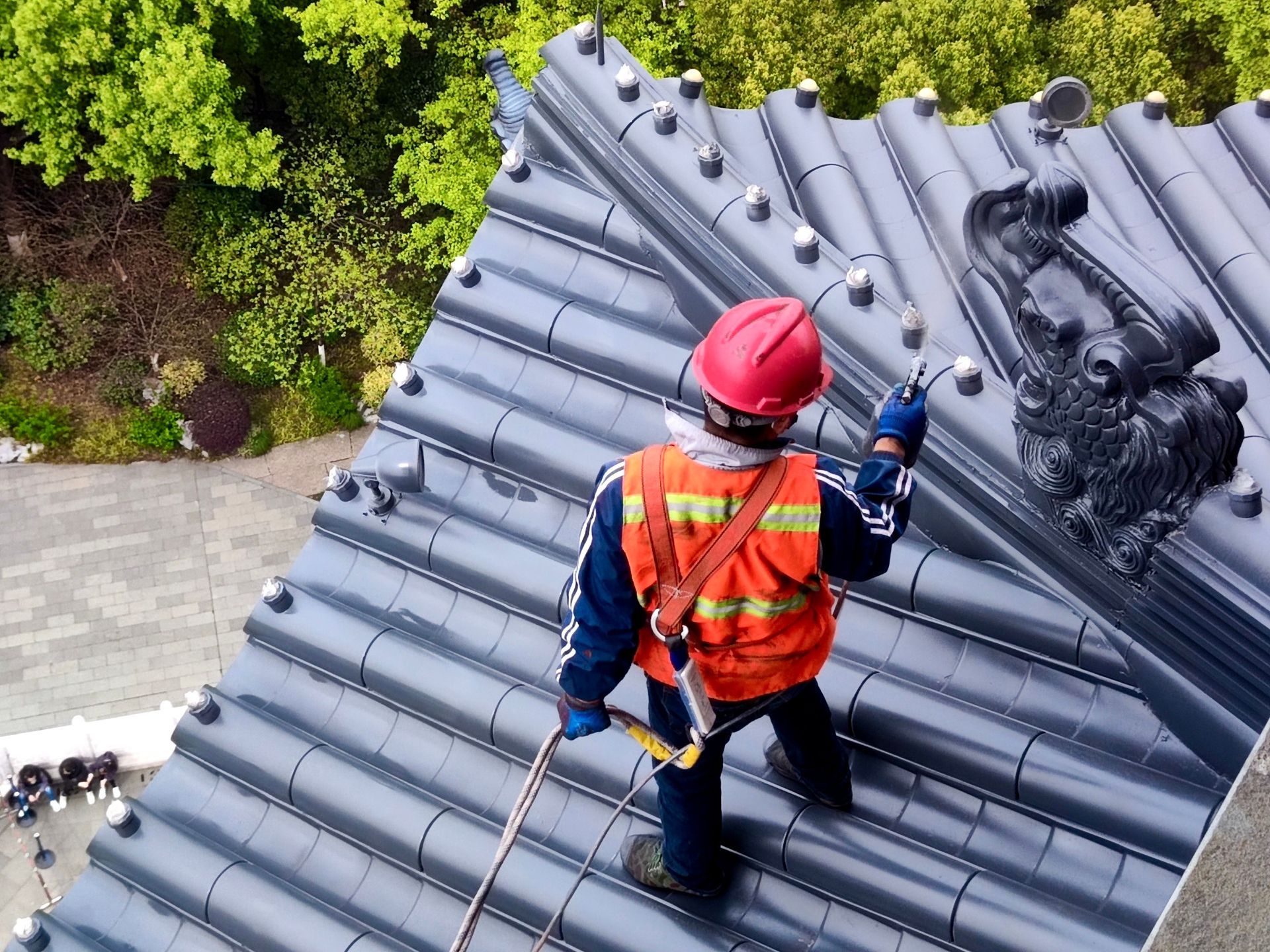 Worker in red helmet and orange vest rappels on a gray roof, cleaning near a tree-lined yard.