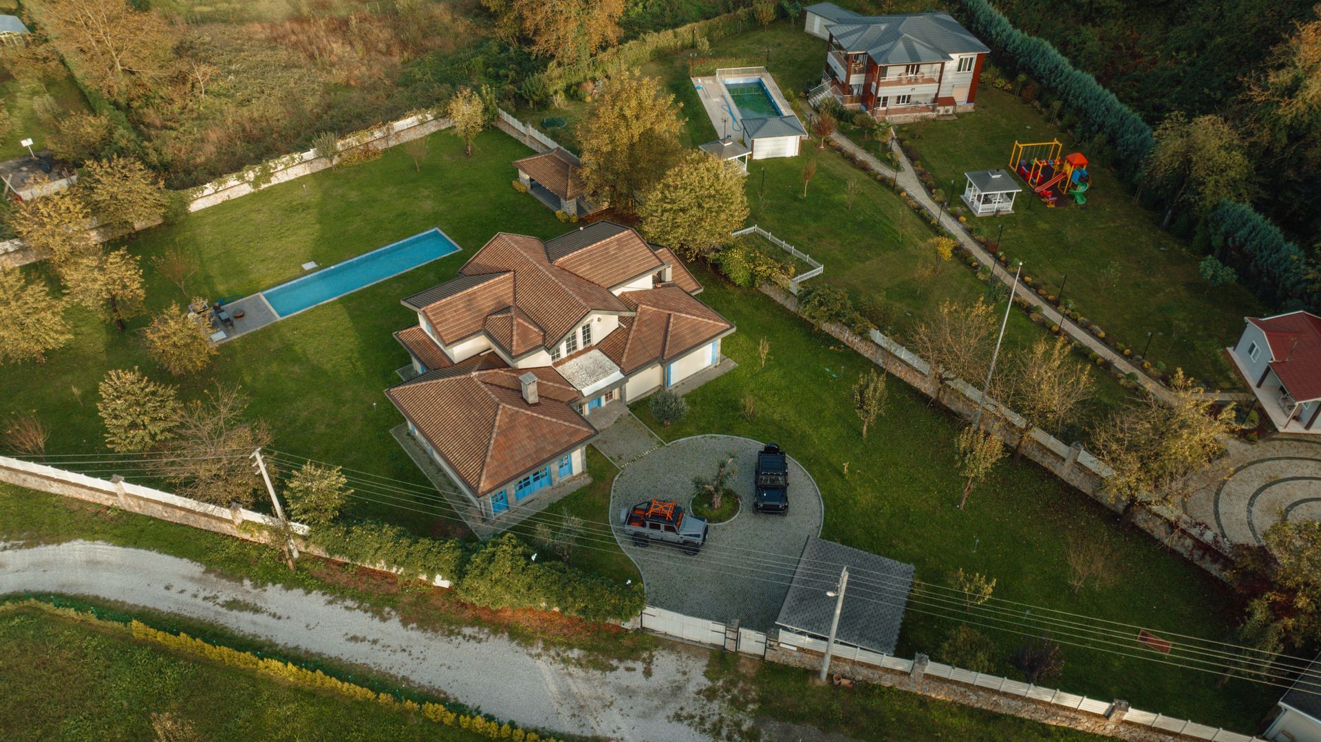 Aerial view of a house with a red-tiled roof, driveway, and surrounding greenery by a road.