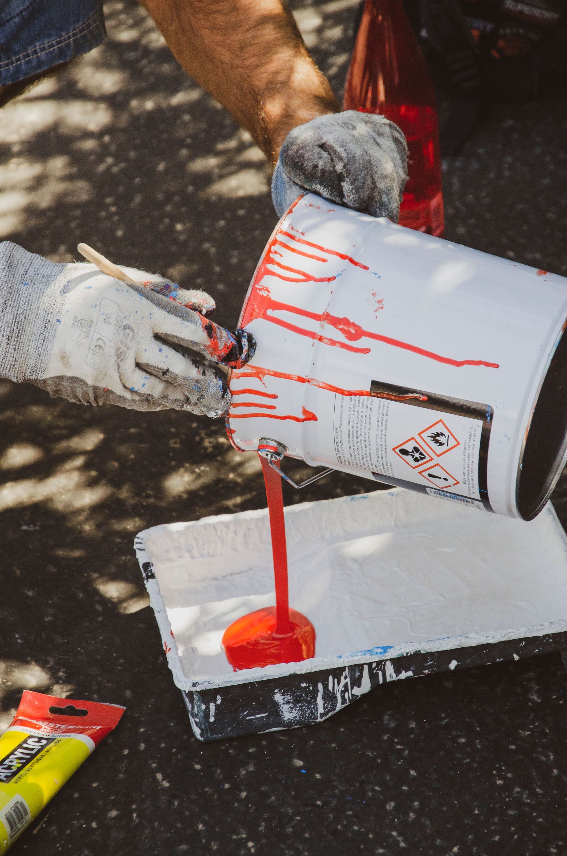 Gloved hands pour bright red paint from a white can onto a white board outdoors.