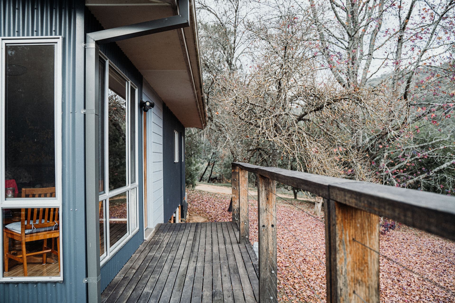 Wooden deck walkway beside a house, with railing and trees beyond a glass door.