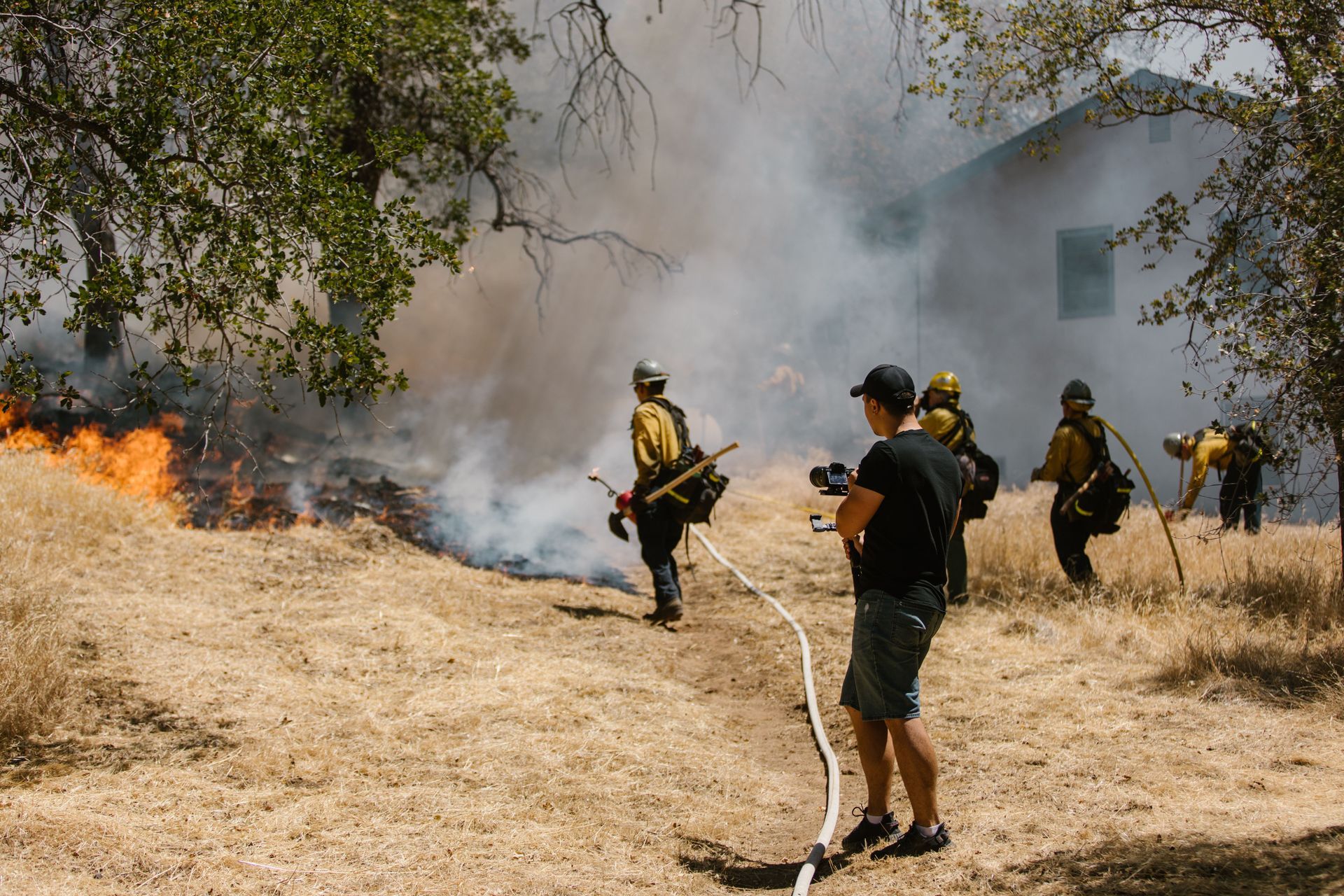 Firefighters battling a brush fire near a house, with smoke and dry grass under trees