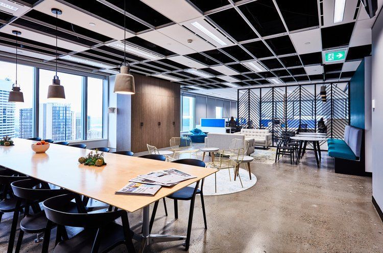 Modern office lounge with tables, chairs, pendant lights, and city views through large windows
