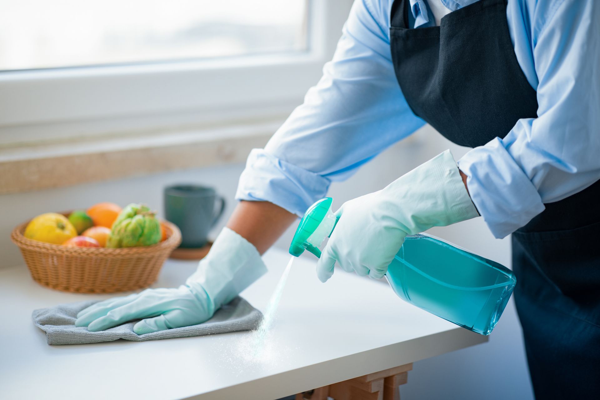 Persona limpiando encimera blanca con botella de spray, usando guantes y delantal.