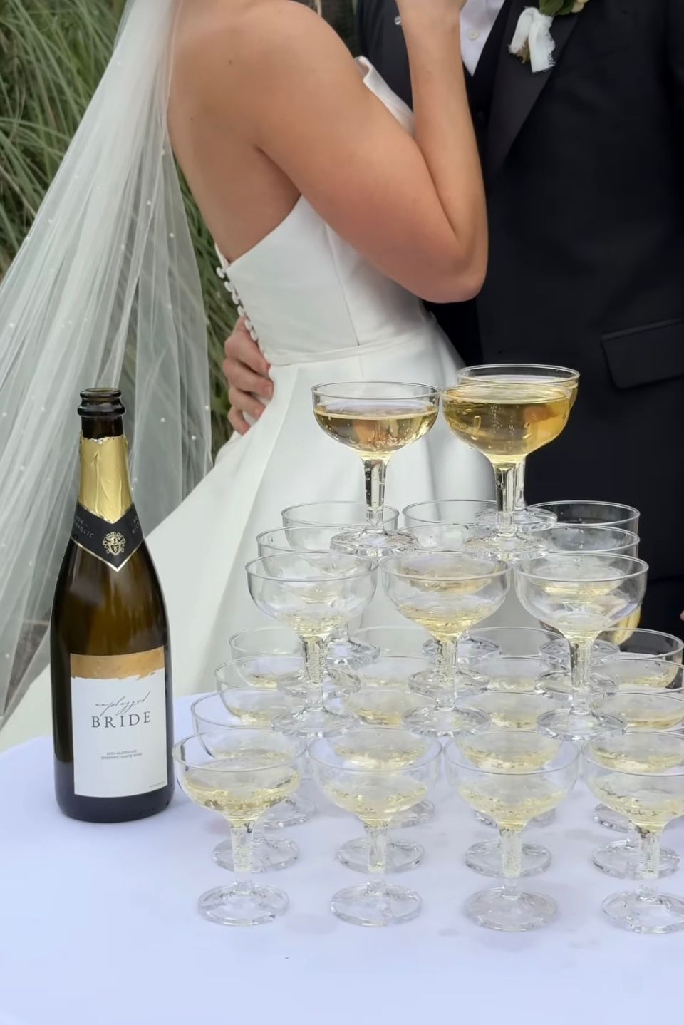 A bride and groom are standing next to a bottle of champagne and glasses of champagne.