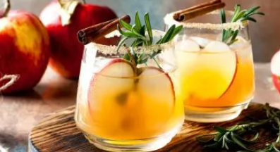 Two glasses of apple cider with cinnamon sticks and rosemary on a wooden cutting board.
