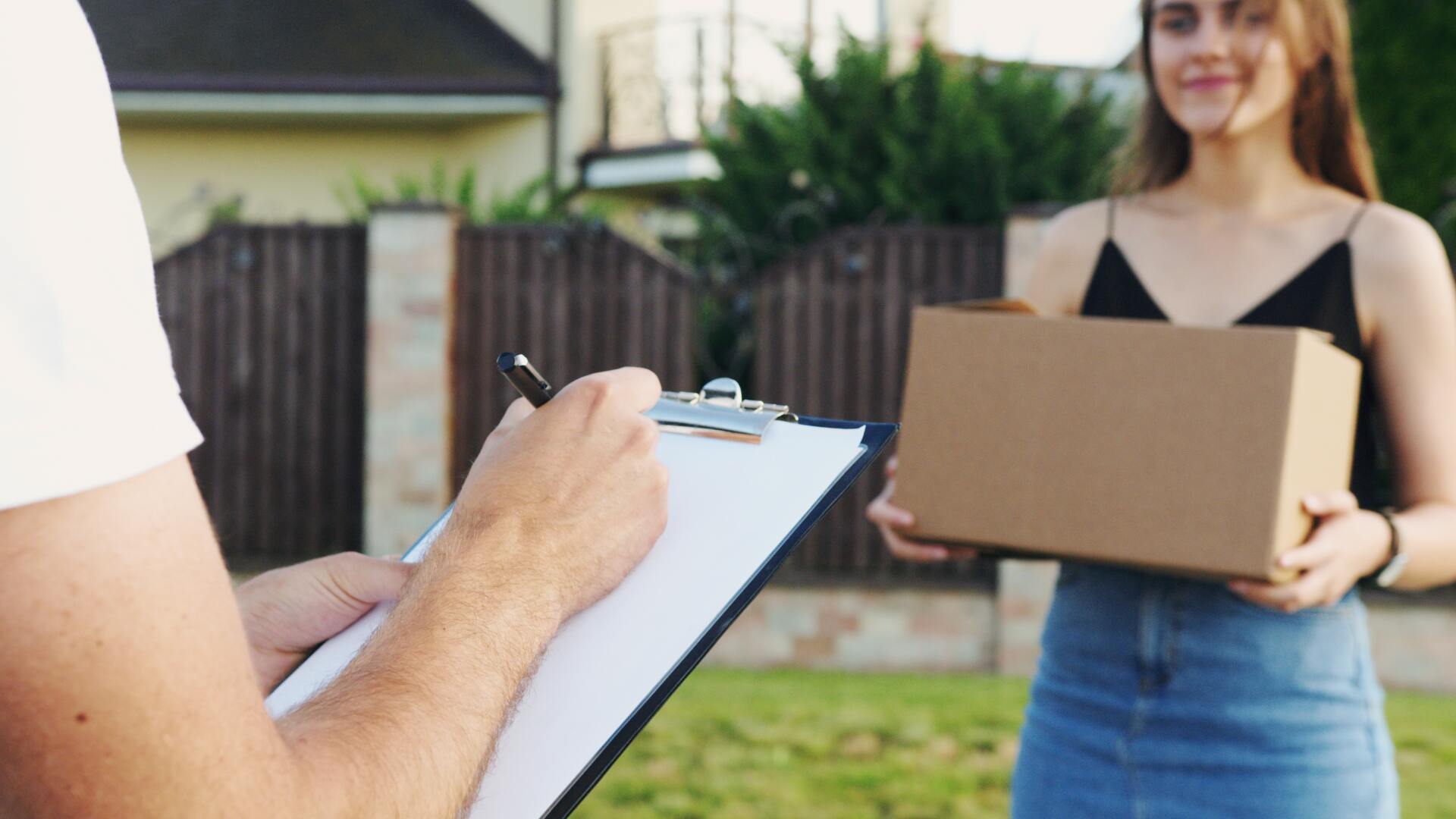 A woman is holding a cardboard box while a delivery man writes on a clipboard.