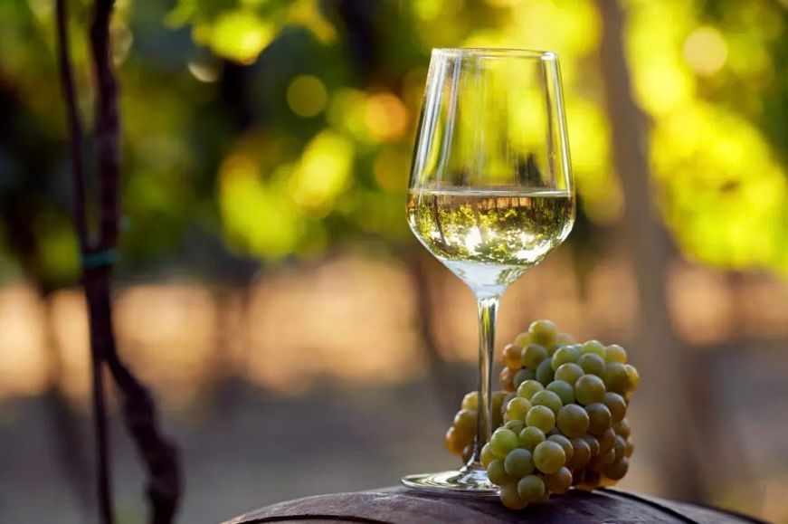 A glass of white wine is sitting on top of a wooden barrel next to a bunch of grapes.