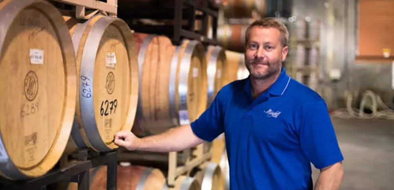 A man in a blue shirt is standing in front of a row of wine barrels.
