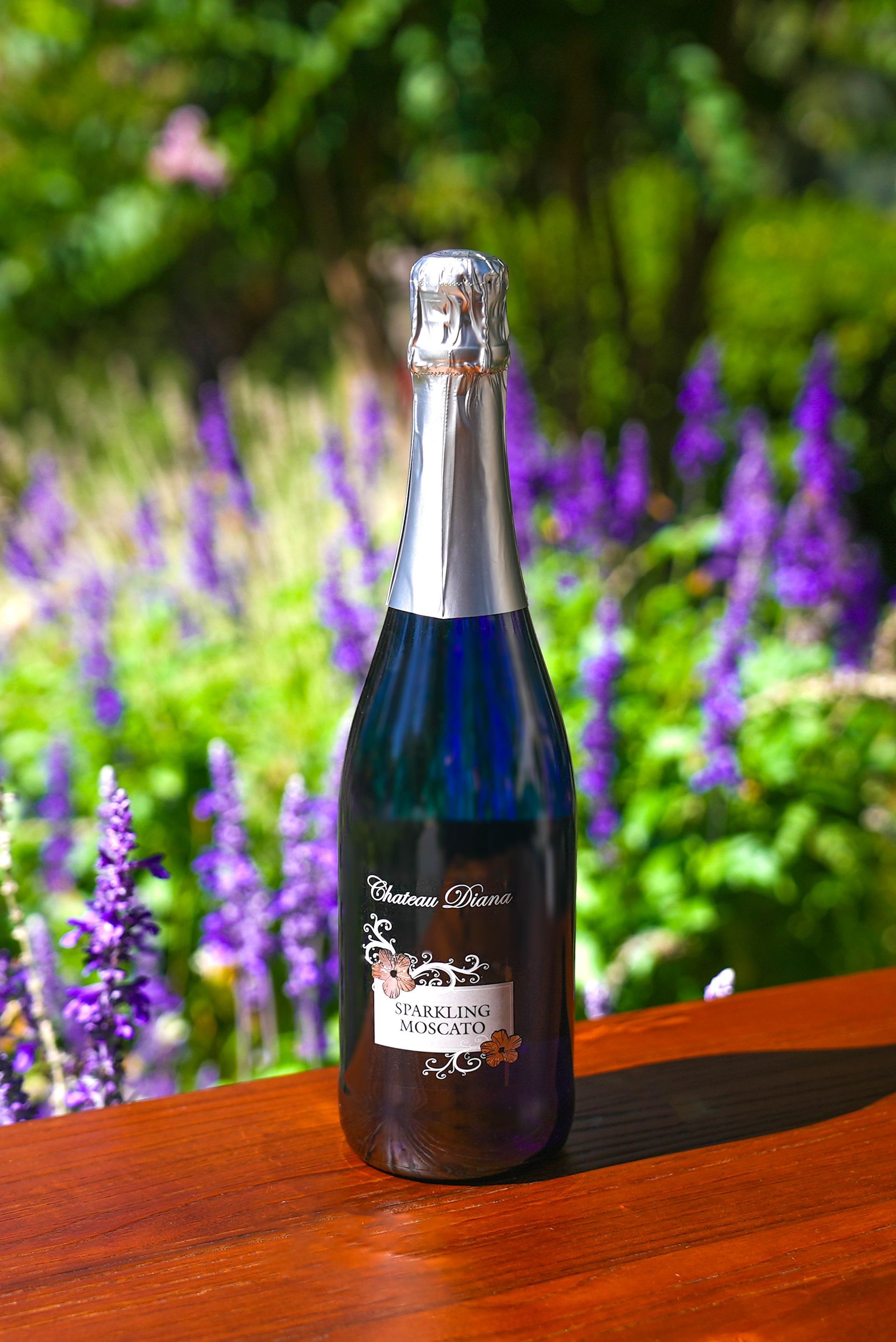 A bottle of blue champagne is sitting on a wooden table in front of purple flowers.