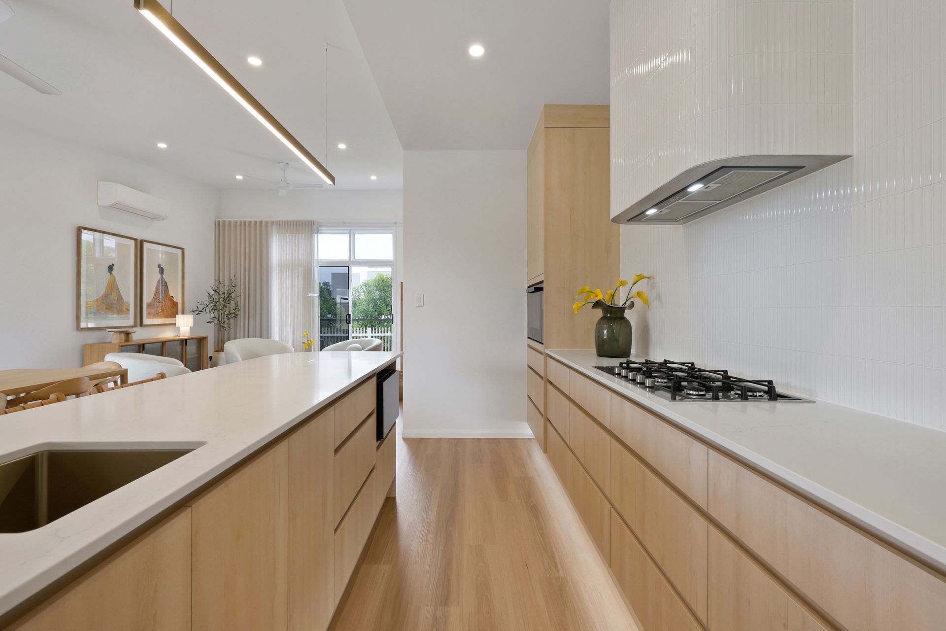 A contemporary kitchen featuring floor-to-ceiling white upper cabinets, slate-grey lower drawers with gold timber pulls, a stainless steel gas cooktop, vertical wood-look tile splashback, open timber shelving with decorative vases, a wall oven, and a walk-in butler's pantry with a coffee machine. Light grey floor tiles and a window with tropical garden views complete the space.