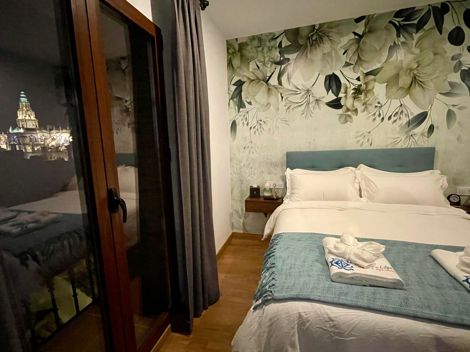 Dormitorio con papel tapiz floral, una cama con detalles azules y una vista de la ciudad visible en el reflejo de la puerta.