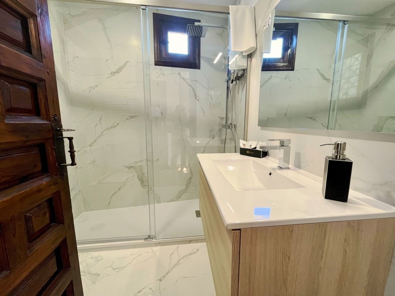 Baño con paredes efecto mármol, tocador de madera, ducha de cristal y puerta de madera color marrón.