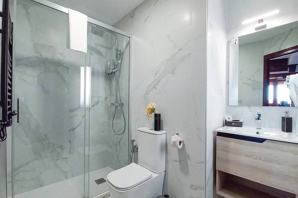Baño moderno con paredes de mármol blanco, ducha de cristal y tocador de madera.
