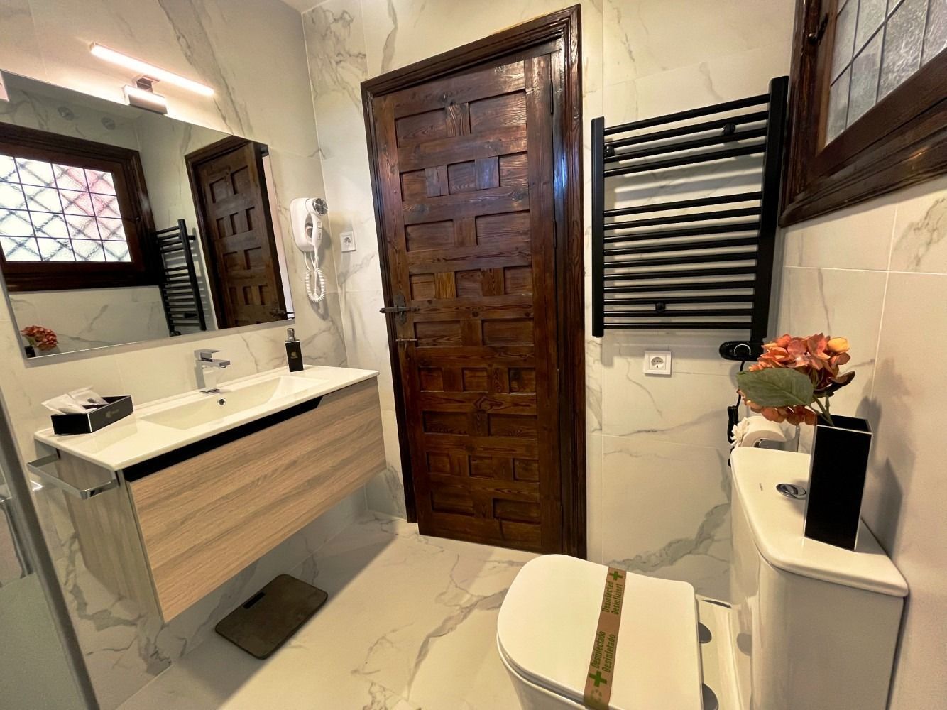 Baño con azulejos imitación mármol, puerta y tocador de madera, inodoro blanco, calentador de toallas negro.