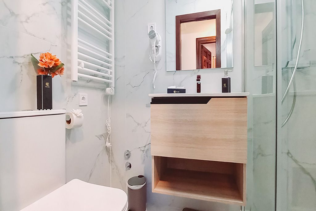 Baño con paredes de mármol blanco, tocador de madera, inodoro y ducha.