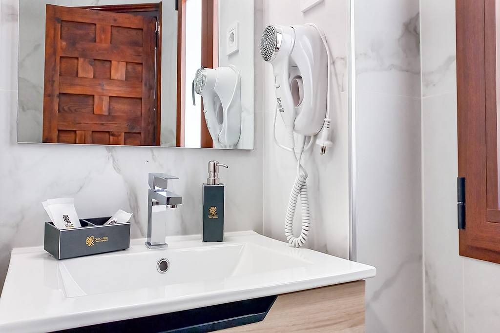 Baño con lavabo blanco, espejo y secador de pelo montado en la pared. Puerta y ventana de madera a los lados.