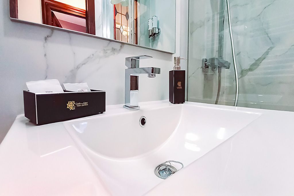 Lavabo blanco con grifo cromado, caja de pañuelos marrón y frasco de perfume en un baño moderno.