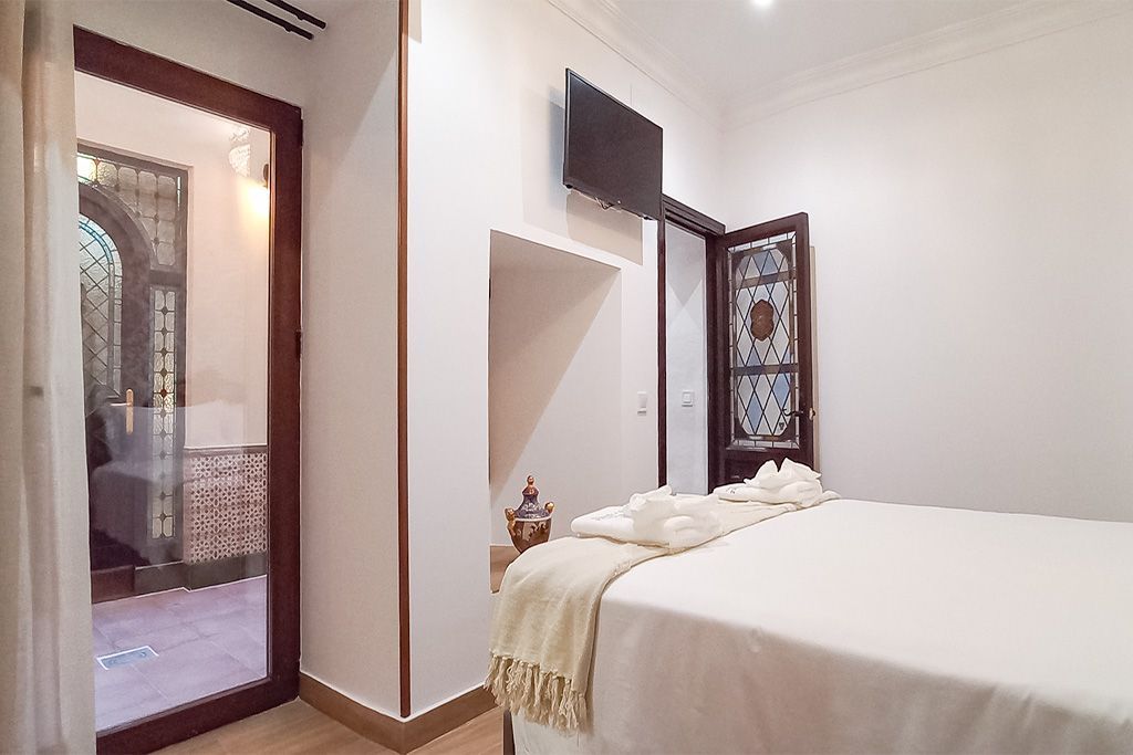 Dormitorio con ropa de cama blanca, televisor en la pared, puerta del baño visible.