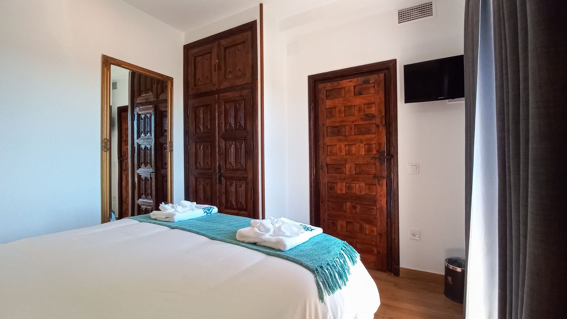 Dormitorio con cama blanca, manta turquesa, puertas y armario de madera y un pequeño televisor.
