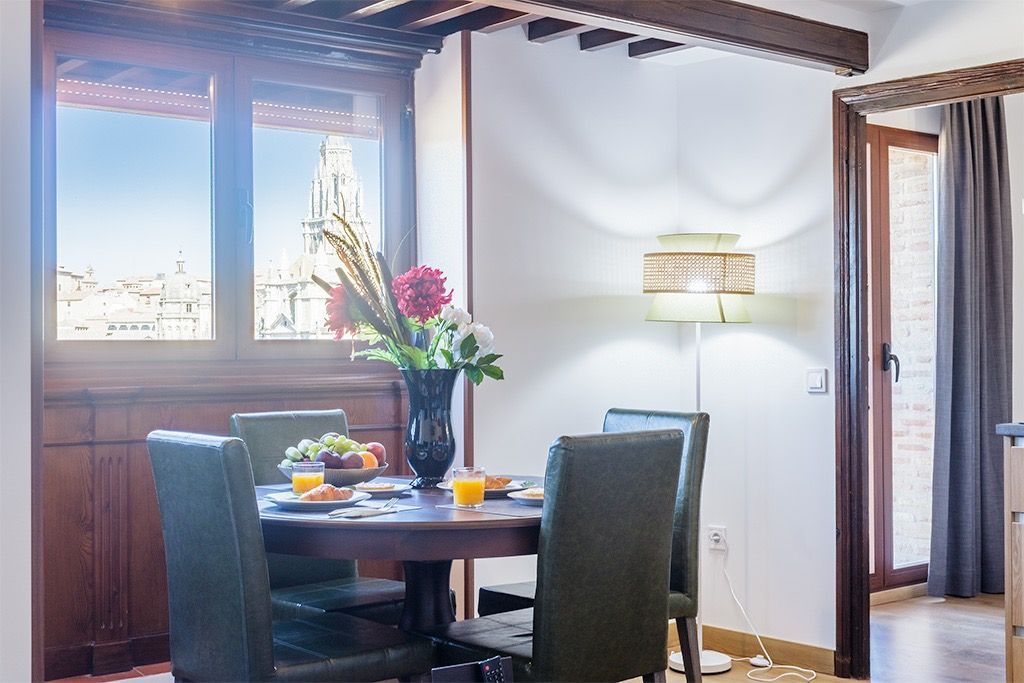 Comedor con mesa puesta para desayuno, flores, vista a la ciudad desde la ventana, luz natural.