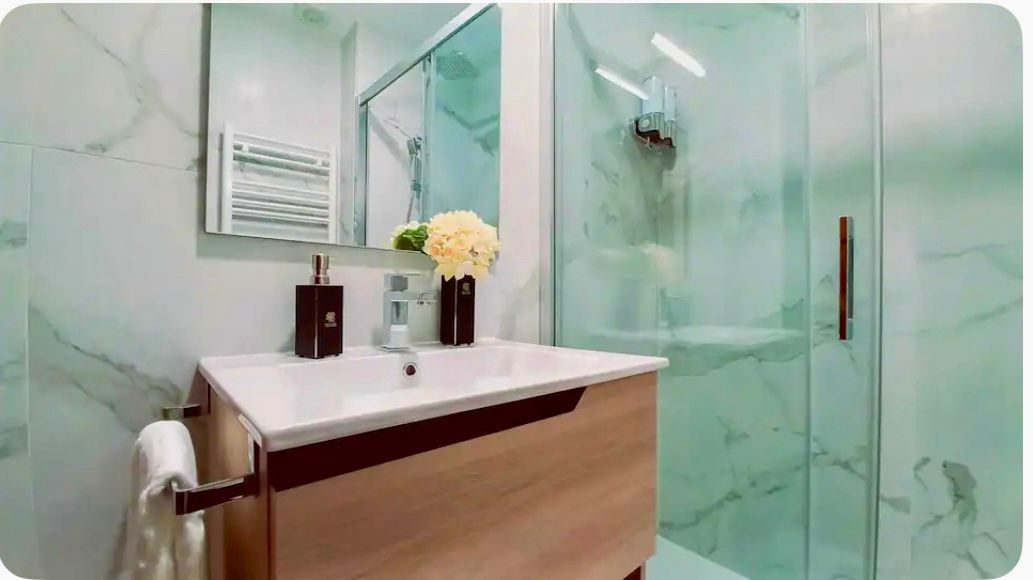 Baño con tocador de madera, lavabo blanco y ducha de cristal. Paredes con motivos de mármol, espejo y toallero.