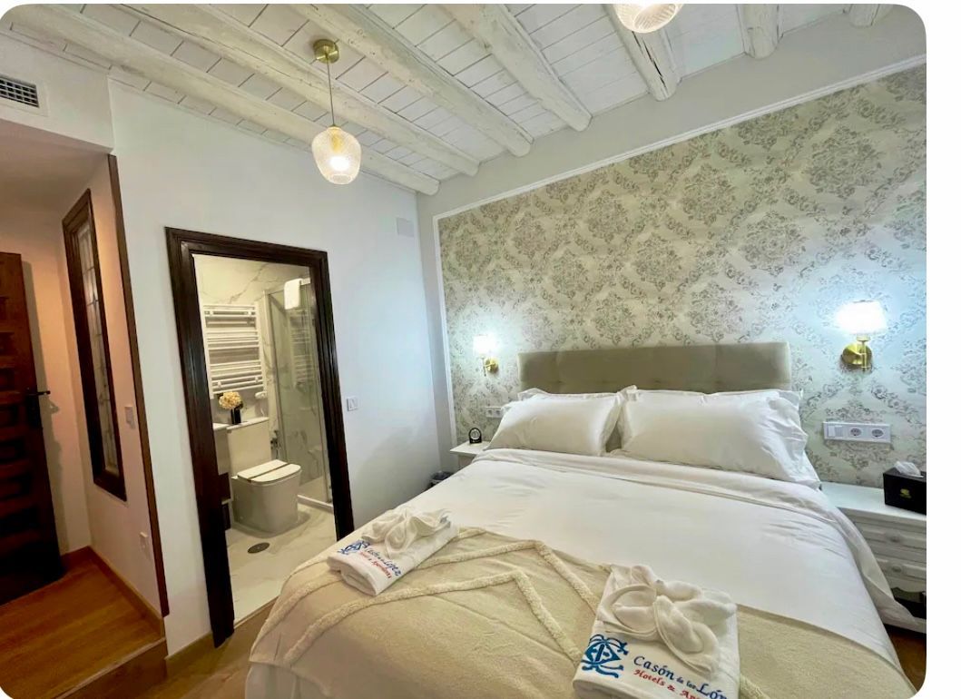 Dormitorio con cama de matrimonio, baño a la vista, papel pintado, sábanas blancas y techo decorativo.