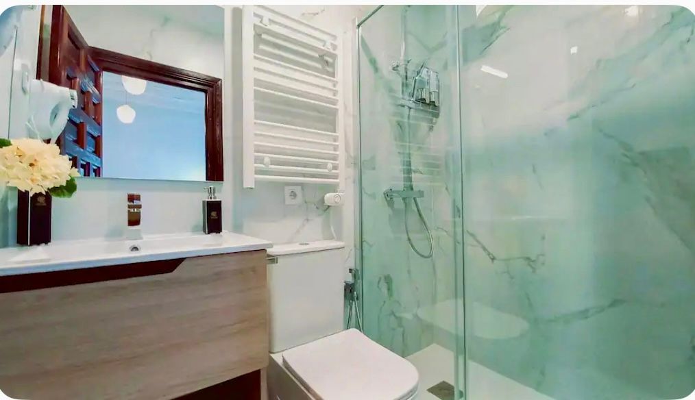 Baño con azulejos blancos y mármol, tocador de madera, ducha e inodoro blanco.