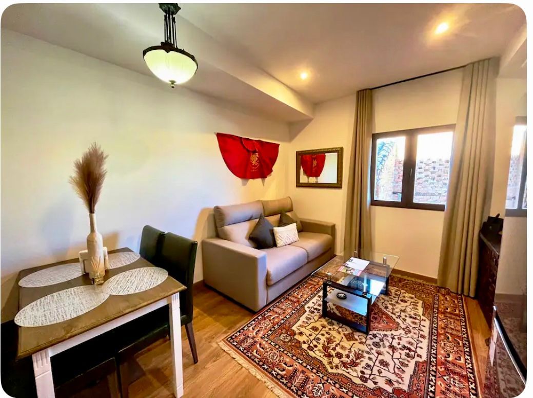 Espacio acogedor: sofá pequeño, alfombra, mesa de comedor y ventana. Tonos cálidos, paredes neutras y detalles decorativos.