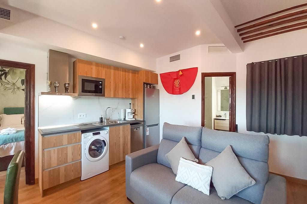 Pequeño apartamento diáfano: cocina con electrodomésticos, sofá, cama vista, suelos de madera, paredes blancas.