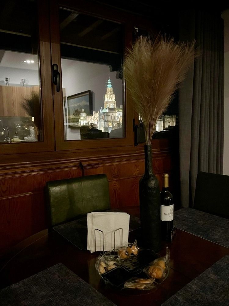 Vista interior, mostrando una ventana con vista a la ciudad, mesa, sillas, botella y plantas decorativas.