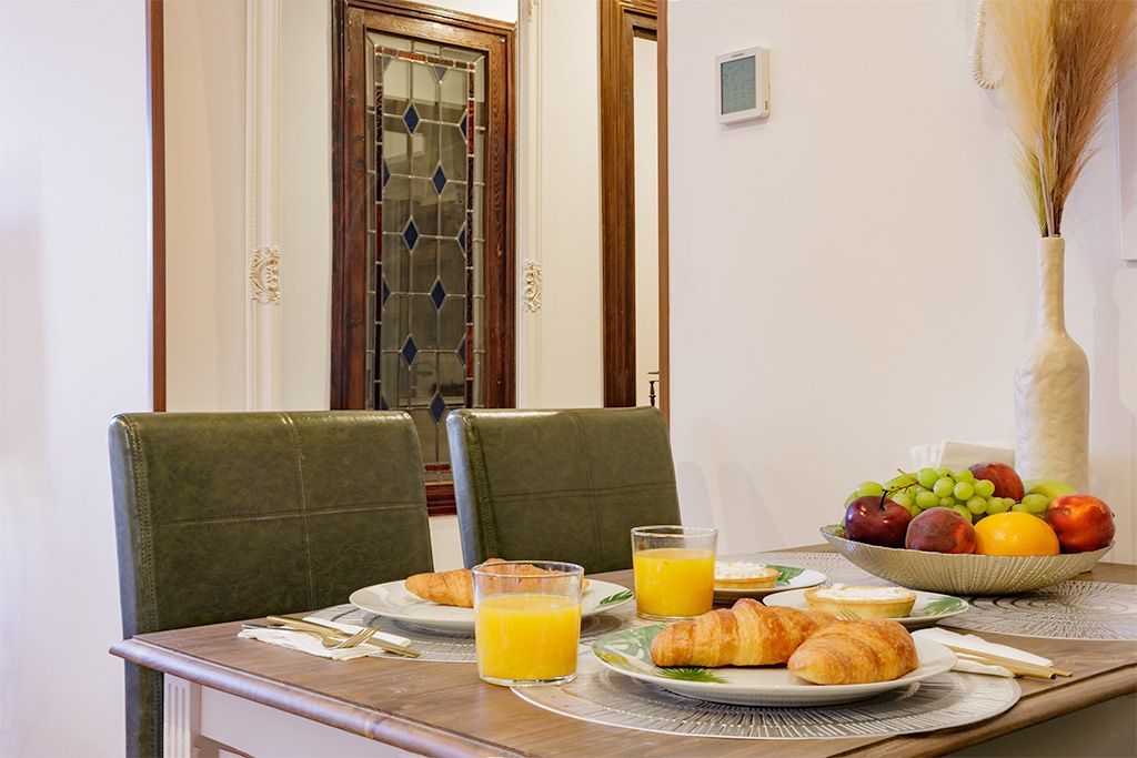 Mesa de desayuno preparada para dos con croissants, jugo, fruta y sillas verdes.