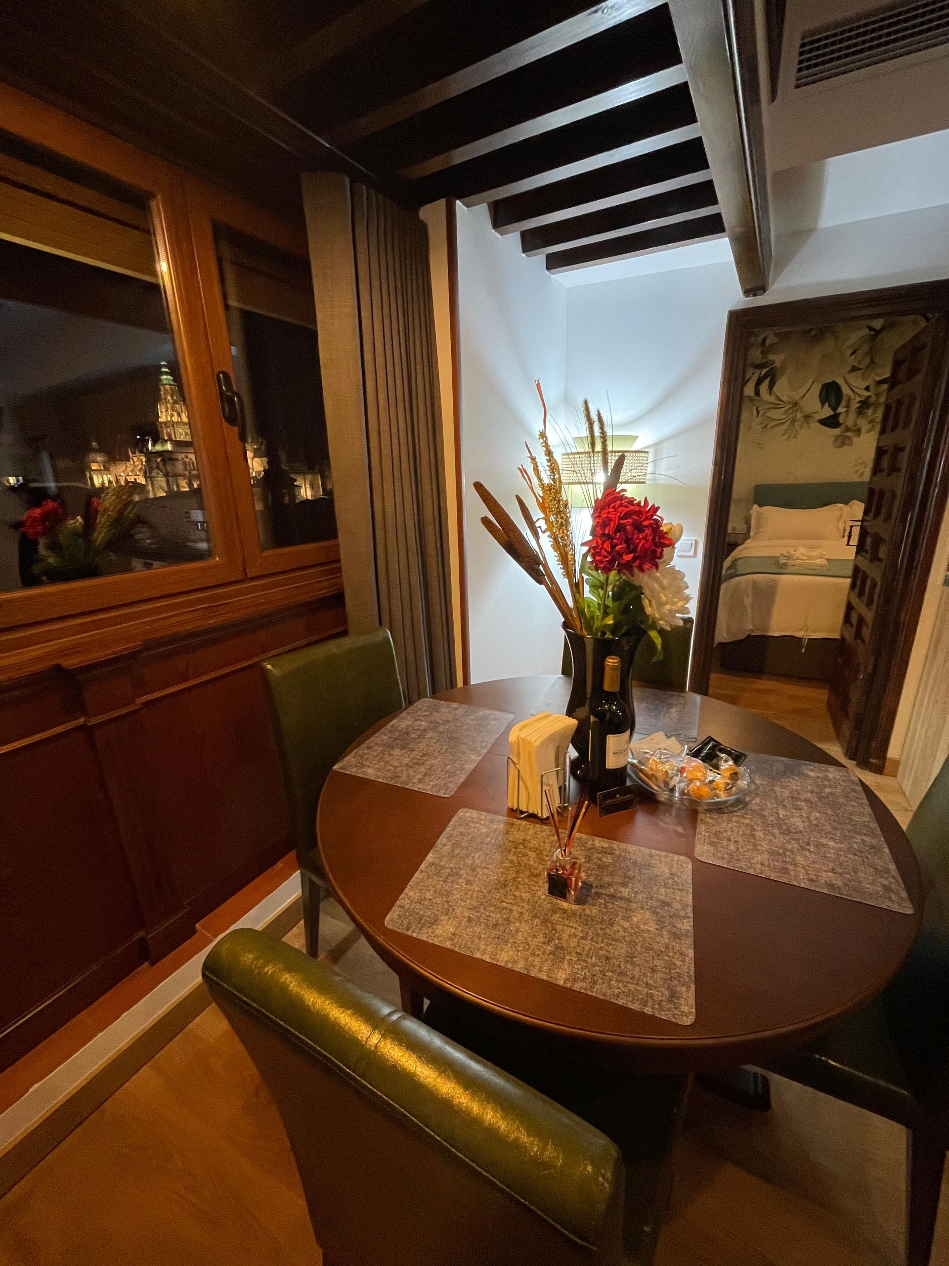 Acogedor comedor con mesa redonda de madera para dos. Sillas de cuero verde, flores en un jarrón y la puerta del dormitorio a la vista.
