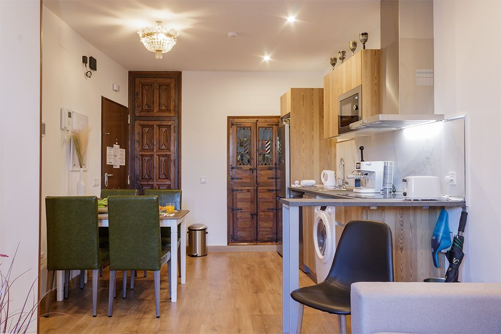 Fotografía interior de una sala de estar con cocina y comedor. Suelos de madera, sillas verdes y puertas antiguas.