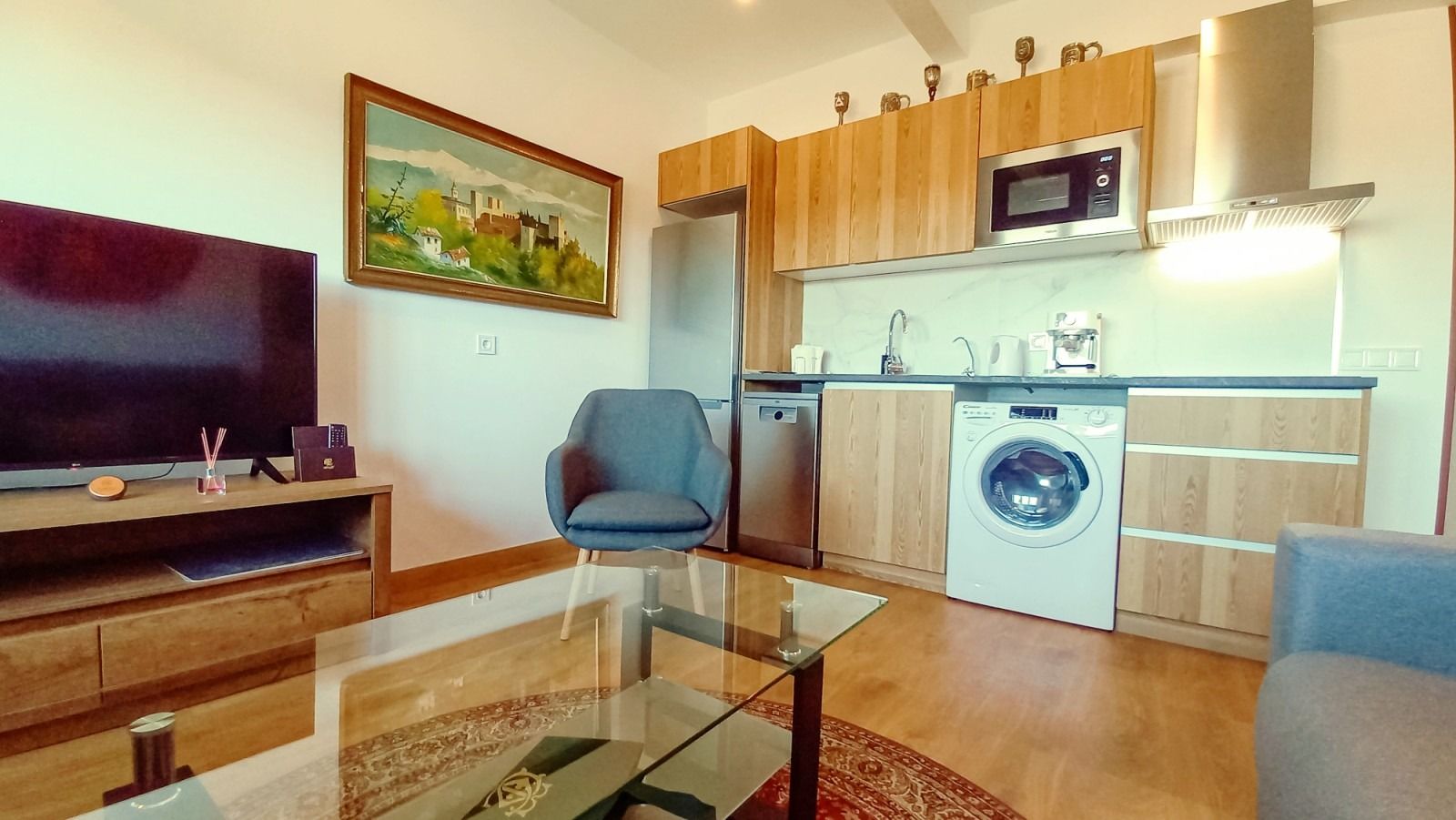 Pequeño apartamento con TV, cocina (microondas, nevera, lavadora), silla azul, armarios de madera y mesa de centro de cristal.