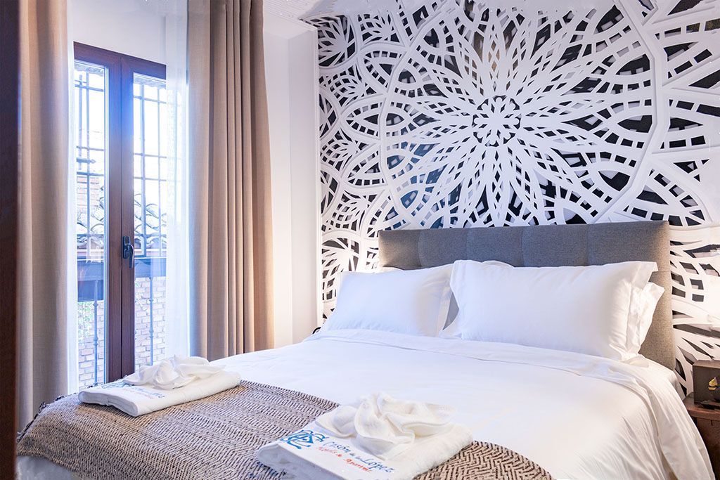 Dormitorio con cama blanca, pared estampada y ventana. Cortinas y mesita de noche en beige claro.
