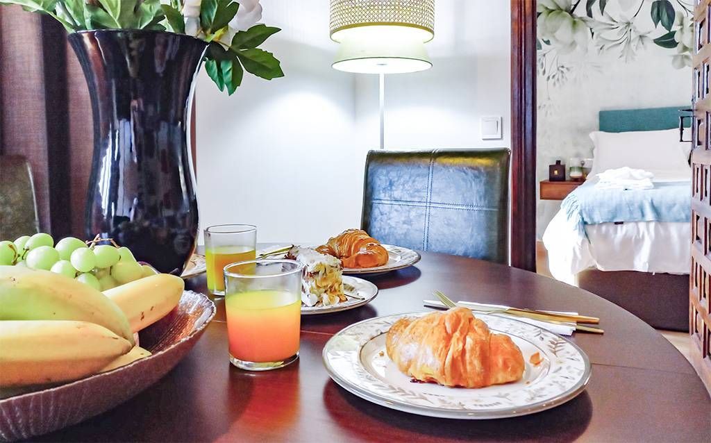 Mesa de desayuno con croissant, fruta, jugo y vista del dormitorio a través de la puerta.