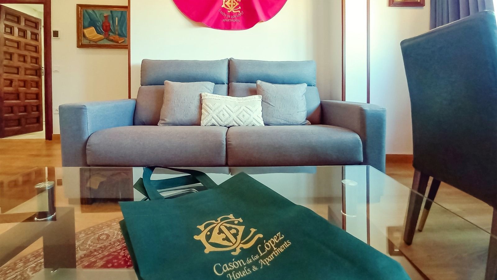 Sala de estar con sofá azul, mesa de cristal y bolso verde. Obra de arte rosa, cuadro y puerta de madera al fondo.