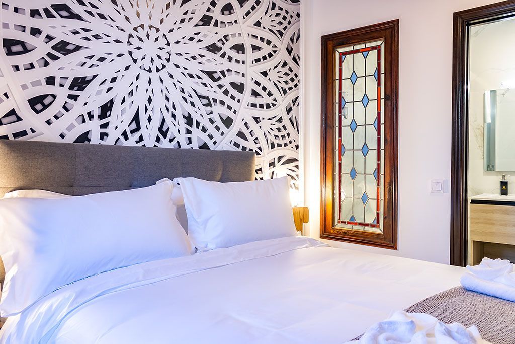 Dormitorio con ropa de cama blanca, pared estampada, vidriera y puerta de baño abierta.