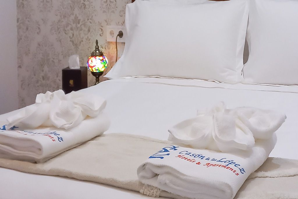 Cama de hotel con sábanas blancas, toallas dobladas y lámpara de colores.