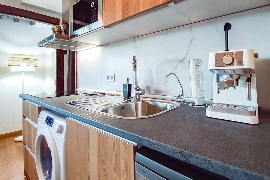 Cocina con gabinetes de madera, fregadero de acero inoxidable y máquina de café expreso sobre encimera. Lavadora a la vista.